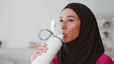 Genç bir Orta Doğulu kadın evde antrenman yaparken bir su şişesiyle su içiyor. Sağlıklı bir yaşam tarzı ve formda kalma alışkanlığı var..