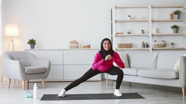 Pembe bluzlu ve tesettürlü genç bir Orta Doğulu kadın oturma odasındaki spor paspasına atlıyor. Konforlu alanda, sağlıklı bir yaşam tarzını destekleyen modern dekorlar ve bitkiler yer alıyor..