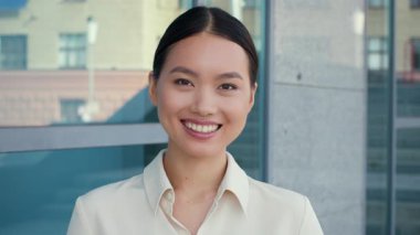 Açık renkli bluz giymiş genç bir Asyalı kadın ofis ortamında duruyor. O, işine bağlanırken kendine güvenen bir gülümsemeye sahiptir. Güneş ışığı modern mekanı aydınlatır..