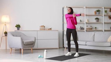 Modern bir oturma odasında, tesettürlü genç bir Orta Doğulu kadın antrenmanına hazırlanmak için kollarını uzatıyor. Ev ortamında spor ve sağlıklı bir yaşam tarzını benimsiyor..