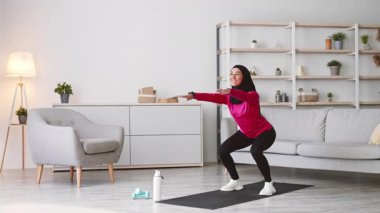 Orta Doğulu tesettürlü genç bir kadın oturma odasında çömeliyor. Etrafı spor aletleriyle çevrili. Egzersizine odaklanıyor, sağlıklı bir yaşam tarzını ve spor programını destekliyor..