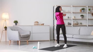 Türbanlı genç bir kadın şık oturma odasında egzersiz yapıyor. Formda kalırken sağlıklı bir yaşam tarzını geliştiriyor. Odaklanmış ve enerjik, egzersizin tadını çıkarıyor..