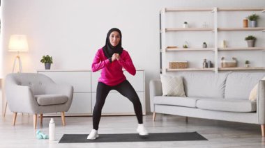 Genç bir Orta Doğulu kadın evde bir antrenmana katılıyor. Konforlu bir oturma odasında spor yaparken azim ve odaklanma gösteriyor..