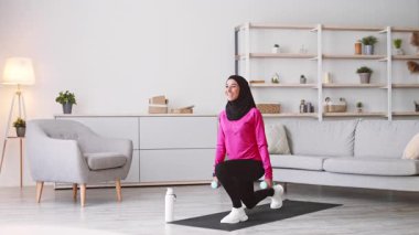 Başörtüsü takan genç bir Orta Doğulu kadın oturma odasına atlıyor, ağırlıklar ve su şişesiyle egzersiz yaparken sağlıklı bir yaşam tarzına olan bağlılığını gösteriyor..