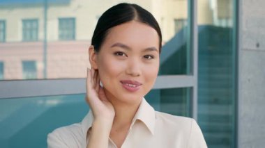 Asyalı genç bir iş kadını cam bir duvarın önünde kendine güvenerek duruyor, profesyonellik ve soğukkanlılık yayıyor..