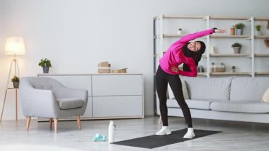 Genç bir Orta Doğulu kadın oturma odasında etek giyerken esneme egzersizleri yapıyor. Ev ortamında egzersiz yaparak sağlıklı bir yaşam tarzı sürdürüyor..