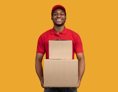 Teslimat servisi konsepti. Kırmızı şapkalı ve üniformalı gülümseyen Afro-Amerikan erkek kurye elinde karton kutular, paketlerle turuncu stüdyo arka planında tek başına duruyor.