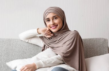 Modern Modesty. Portrait Of Beautiful Arabic Girl In Hijab Sitting On Couch At Home And Smiling To Camera, Posing On Sofa, Free Space