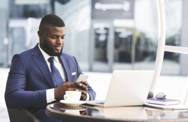 İş hayatı tarzı. Gülümseyen İşadamı Cep Telefonu Mesajını Kullanıyor Laptop 'ta çalışıyor Şehir Bölgesi' ndeki açık kafede oturuyor