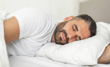 Sakallı bir adam yatakta yan yatmış, mışıl mışıl uyuyor. Gözleri kapalı ve yüzünde rahatlamış bir ifade var. O beyaz bir tişört giyiyor ve beyaz bir battaniyeyle kaplı..