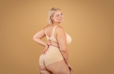 Vücut pozitiftir. Güzel kıvrımlı kadın bej renkli stüdyo arka planında poz veriyor, çekici artı iç çamaşırlı sarışın yarı dönüşmüş ve kameraya gülümsüyor, boşluğu kopyalıyor.