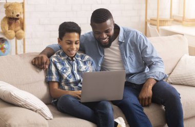 Caring Father Teaching Son To Use Laptop, Sitting On Couch At Home Together