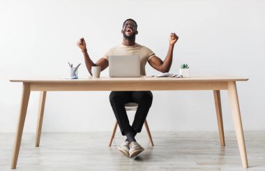 Heyecanlı bin yıllık siyahi bir adamın diz üstü bilgisayarın önünde EVET işareti yapan, başarıyı ya da başarıyı kutlayan, merkez büroda masa başında oturan, fotokopi çeken uzun bir portresi. Uzak iş, çevrimiçi kazanma kavramı