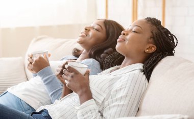 İki siyah kız arkadaş kanepede kahve içip dinleniyor, gözleri kapalı yatıyor ve zor bir günün ardından dinleniyor.