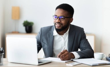 Yakışıklı siyah girişimci dizüstü bilgisayar kullanarak ve modern beyaz ofiste çalışırken notlar alarak, panorama