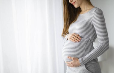 Sıradan kıyafetlerle hamile bir kadını, karnını nazikçe tutarken, beyaz perdeli bir pencerenin yanında dururken gösteren nazik bir görüntü.