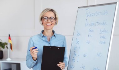 Çevrimiçi eğitim konsepti. Gramer kurallarıyla tahtaya yakın poz veren pozitif Almanca öğretmeninin portresi, kameraya bakıyor ve gülümsüyor, kapalı alanda. Yabancı diller öğretmeni uzaktan ders veriyor