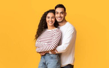 Kameraya sarılan ve gülümseyen genç Arap erkek ve kadının portresi, sarı arka planda birlikte poz verirken romantik Orta Doğu çifti,