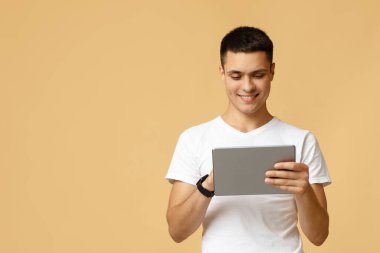 Çalışmak, eğlenmek, dinlenmek ve iletişim kurmak için bir cihaz. Mutlu genç adam arkadaşlarına yazmak için tablet kullanır, oyun oynar ve bej arka planda izole edilmiş sohbet eder, stüdyo çekimi, boş alan