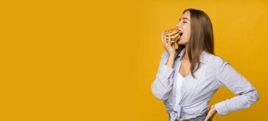 Hile yemeği. Aç milenyum kızı hamburger yiyor sağlıksız fast food yiyor sarı stüdyo arka planında duruyor. Panorama, boşluk