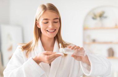 Bornozlu genç bayan pamuk pedi ve bir şişe yüz losyonu kullanıyor. Banyodaki tonik veya mikiler suyuyla deriyi nemlendirip temizliyor. Yüzü kozmetik ürünlerle nemlendirmeye hazır bir kadın.