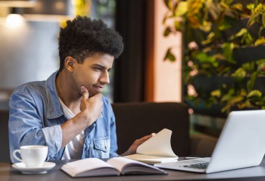 Konsantre Afrikalı genç öğrenci ev ödevi yapıyor, kafede dizüstü bilgisayar kullanıyor, çenesini dokunuyor, kopya alanı