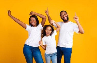 Eğleniyorum. Üç kişilik heyecanlı zenci bir ailenin portresi dans ediyor ve oynaşıyor. Mutlu Afrikalı erkek, kadın ve kız sarı stüdyo arka planında favori müziğin tadını çıkarıyorlar.