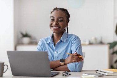 Öğrenciler için çalışmak. Genç siyahi bayan ofis çalışanı pozu dizüstü bilgisayarla masada oturuyor ve kameraya gülümsüyor genç stajyer yeni işin tadını çıkarıyor, boşluğu kopyalıyor.