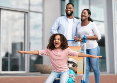 Yaz Tatili Konsepti. Neşeli, pozitif, siyah bir kızın el pençe divan uçmaya hazır, bagaj vagonunda oturduğu portresi. Aile yük arabasıyla yürüyor, kızına biniyor.