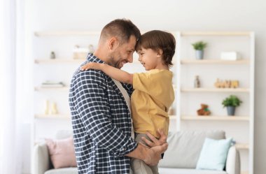 Baba oğul ilişkisi. Mutlu baba küçük oğlunu elinde tutuyor, alınları birbirine bağlanıyor ve gülümsüyor, evde iç kısımda duruyor.