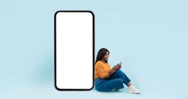 Neşeli genç siyahi kadın cep telefonu kullanıyor. Dev akıllı telefonun yanında oturuyor. Mavi ekranlı, mavi stüdyo arka planda, tüm vücut uzunluğunda. Web sitesi uygulaması için modelleme, reklam için ücretsiz kopya alanı