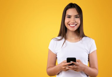 Halk ve Teknoloji Konsepti. Portakal rengi stüdyo arka planında izole edilmiş akıllı telefon kullanan sıradan gülümseyen genç kadın portresi kameraya bakıyor. İnternette sohbet eden kadın, sosyal medyada gezinen.