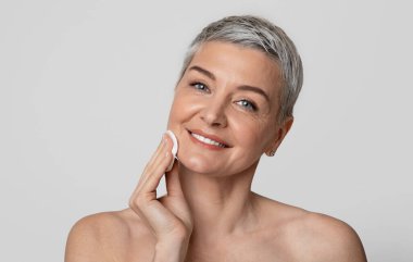 Cilt bakımı. Pamuk pedi kullanan, makyajdan deriyi temizleyen ve kameraya bakan, arka planda duran güzel, olgun bir kadın..