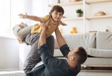 Genç, mutlu, beyaz bir baba yerde yatıyor ve kucağında küçük oğlunu taşıyor. Çocuk uçakla oynuyor ve kameraya gülümsüyor. Komik aile aktiviteleri kavramı.