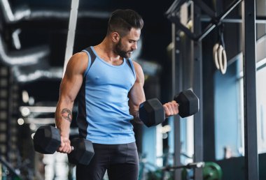 Güçlü, kaslı bir adam halterlerle egzersiz yapıyor, spor salonunda çalışıyor, boş yer var.