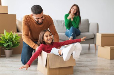 Hareket halindeyken eğlenmek. Mutlu Arap aile portresi yeni bir dairede birlikte eğleniyor, paketleri açıyor, gülümseyen baba heyecanlı küçük kızı oturma odasında karton bir kutuda gezdiriyor, kanepede oturan kadın.