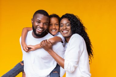 Aile sevgisi. Mutlu Afrikalı Amerikalı ebeveynler, küçük kızlarıyla poz veriyorlar, birlikte gülüyorlar ve sarı arka plandan kameraya bakıyorlar.