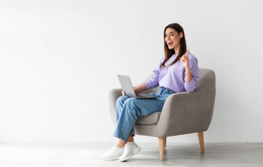 Bin yıllık beyaz bir kadının yaratıcı bir fikri var, Eureka 'yı işaret ediyor, dizüstü bilgisayarla beyaz stüdyo duvarına yaslanmış bir koltukta oturuyor, kopyalama alanı. Soruna çözüm bulan genç bayan.