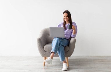 İnternette çalışan genç beyaz bir kadın, elinde kahve, koltuğunda oturuyor ve beyaz stüdyo duvarına karşı dizüstü bilgisayar kullanıyor. Taşınabilir bilgisayarda internette sörf yapan neşeli milenyum kadını.