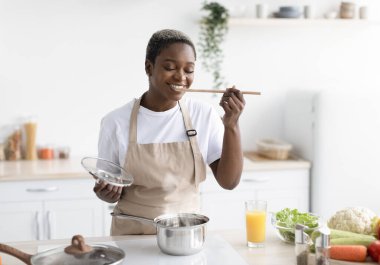 Gülümseyen bin yıllık Afro-Amerikan kadın önlüklü evde yemek pişiriyor ve mutfağın içinde yemek tadıyor. Öğle yemeği, ev hanımı ve hobi hazırlayın, covid-19 salgını sırasında sağlık hizmeti, kopyalama alanı
