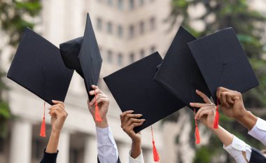 Mezuniyet konsepti. Çok ırklı bir grup öğrenci şapkalarla el kaldırıyor. Öğrencilerin yetiştirdiği mezuniyet şapkaları ve diplomaları üniversite veya üniversite binalarını devrediyor.