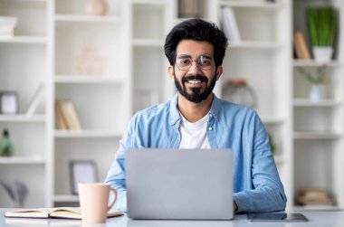 Masasında dizüstü bilgisayarla oturan ve kameraya gülümseyen yakışıklı Hintli genç adam, Millennial Eastern erkek serbest bilgisayar ile çalışıyor, uzak iş fırsatlarının tadını çıkarıyor, Uzay Kopyalama