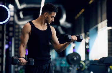 Spor salonunda dambıllı genç Ortadoğulu Erkek Vücut Geliştirici Eğitimi, Modern Spor Kulübü 'nün iç mekânında hafif ağırlıklarla egzersiz yapan kaslı Arap Adam,