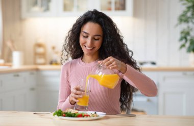 Mutlu bir milenyum kadınının portresi. Mutfakta sağlıklı kahvaltısının tadını çıkarıyor. Neşeli esmer kadın masada oturmuş lezzetli yemekler yiyor ve portakal suyunu sürahide bardağa dolduruyor.