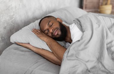 Healthy Sleeping Concept. Portrait of happy young well-slept African American man lying in bed with closed eyes, resting in bedroom on the side in the morning, having hap