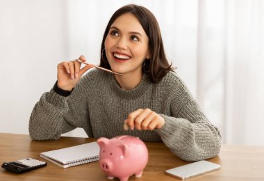 Smiling young Hispanic woman put coin into piggybank save money for vacation. Happy Latin lady manage household budget finances, make investment in piggy bank, dreaming about future