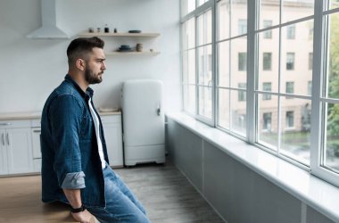 Evde kederli ve depresif bir adamın portresi, covid-19 tecrit, agorafobi, kişisel problemler. Mutsuz bin yıllık Avrupalı yakışıklı adam mutfak içinin geniş penceresine bakıyor.