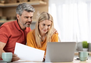 Evde çalışan çekici orta yaşlı eşler, sevecen koca ve eş aile işleri, mutfak masasında dizüstü bilgisayarın önünde oturmak, belgeleri tutmak, kucaklamak, boşluğu kopyalamak