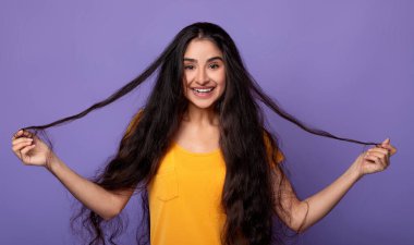 Saç Bakımı ve Terapi Konsepti. Gülümseyen Hintli kadının portresi poz veren ve uzun siyah saçlı ve bukleli. Mor stüdyo arka planında tek başına duran güzel genç bayan, kopyalama alanı