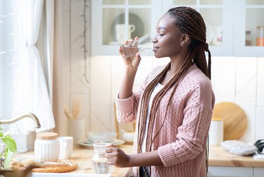 Evdeki mutfaktaki camdan su içen susamış Afrikalı Amerikalı kadın, tezgahın yanında duran ve sağlığını tazeleyen genç siyah kadın, fotokopi alanı ile yan görüş.